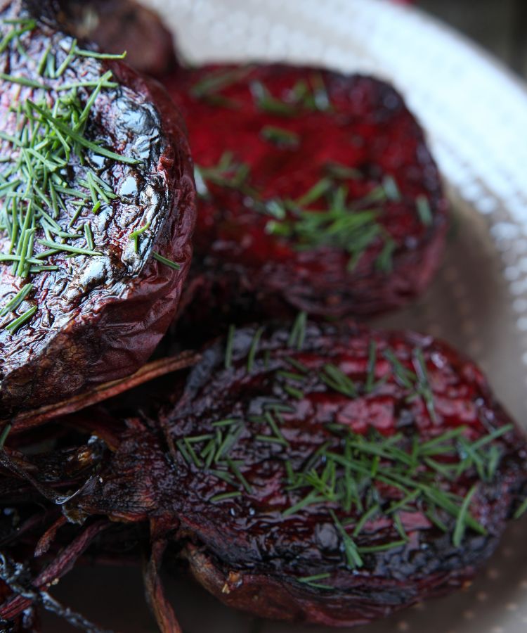 Receitas-Beterraba-3 Beterrabas assadas, cortadas ao meio, em um prato claro. As beterrabas têm uma cor roxo-escura e um brilho caramelizado na superfície, finalizadas com cebolinha picada por cima.