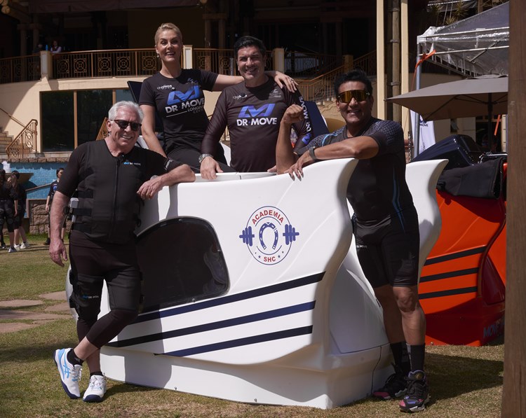 Dr-Move-Longevidade-8 Ana Hickmann e Edu Guedes e outras duas pessoas vestindo roupas de treino escuras e equipamentos de eletroestimulação posam para uma foto em uma área externa. Duas pessoas estão dentro de uma máquina branca com a logo "ACADEMIA SHC" e duas estão do lado de fora.
