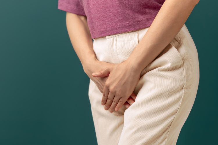 Hpv-Dor-Utero Mulher de pele clara usando blusa rosa, calça bege e colocando as mãos sobre sua genital