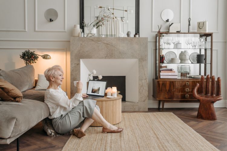 Longevidade-Sentar-Chao Mulher idosa de blusa bege e calça cinza sentada no chão, ao lado de mesa de centro de madeira, em sala com tons beges