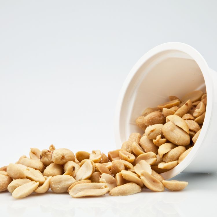 Amendoim-Beneficios-3 A imagem mostra um recipiente branco derramando amendoins sobre uma superfície plana. Os amendoins têm uma coloração que varia do bege ao marrom claro, alguns inteiros e outros partidos. O fundo é neutro e claro, destacando o contraste com os amendoins.