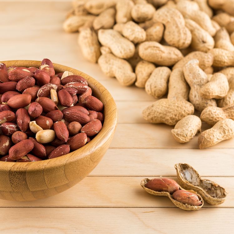 Amendoim-Beneficios-1 A imagem mostra uma tigela de madeira cheia de amendoins descascados e torrados, com alguns amendoins brancos misturados. Ao fundo, há um monte de amendoins com casca. No primeiro plano, um amendoim está aberto.