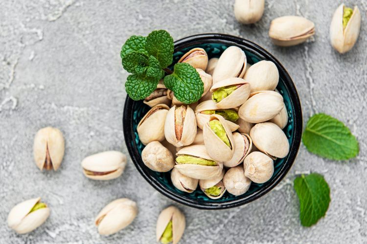 O-Que-E-Pistache-Para-Que-Serve pote preto com noz de pistache dentro e ramo de hortelã em superfície cinza com pistaches e folhas de hortelã