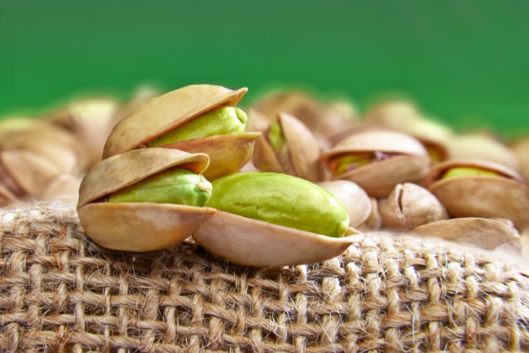 De-Onde-Vem-Pistache 3 nozes bonitas de piscache sob saco de juta com diversas outras nozes de pistache dentro em fundo verde