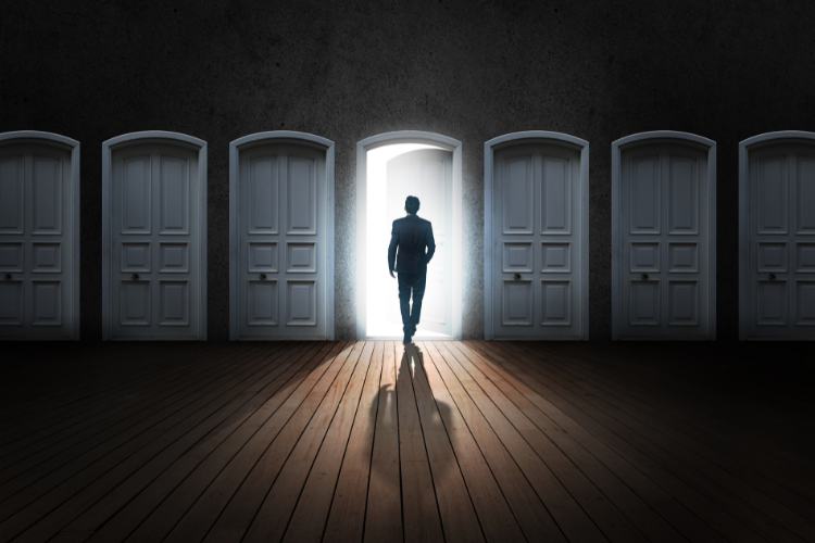 Crise-Em-Oportunidade-1 Homem caminha em direção a uma porta iluminada, enquanto outras permanecem fechadas em um corredor escuro. A imagem simboliza escolhas estratégicas e a capacidade de "transformar crise em oportunidade" ao encontrar novas possibilidades e soluções em momentos difíceis.