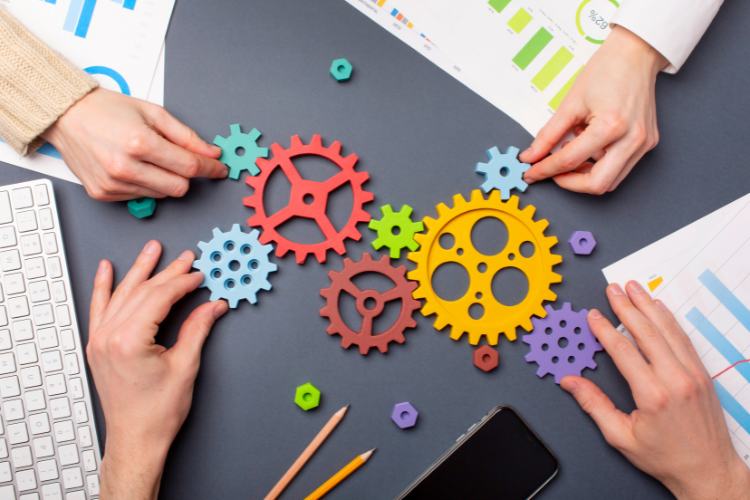 Crise-Em-Oportunidade-10 Quatro mãos ajustam engrenagens coloridas sobre uma mesa com papéis e gráficos, simbolizando trabalho em equipe e inovação. A imagem destaca colaboração estratégica para resolver problemas e "transforme crise em oportunidade" ao encontrar soluções criativas e eficientes.