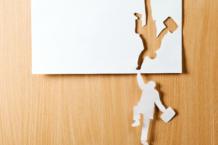 Crise-Em-Oportunidade-2 Recortes de papel formam uma silhueta de pessoa pendurada, segurando outra que parece estar caindo, sobre uma superfície de madeira clara. A imagem simboliza apoio, colaboração, superação de desafios e resiliência em momentos difíceis.