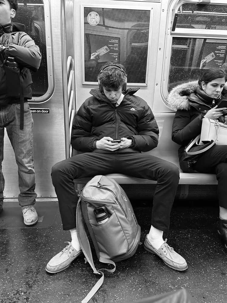 Manspreading-4 Foto em preto e branco de homem usando jaqueta, calça e com mochila no chão ocupando mais de assento. A mulher ao lado dele é obrigada a sentar tomando distância