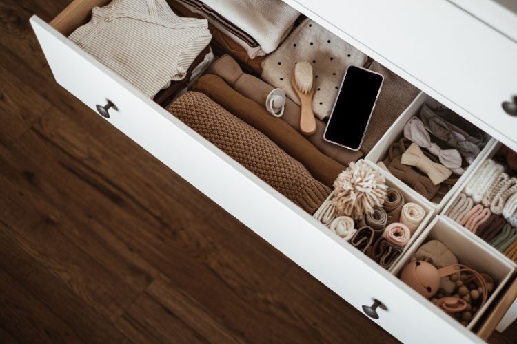 Como-Abandonar-Um-Mau-Habito-2 A imagem mostra uma gaveta aberta, organizada com roupas e acessórios infantis. Há suéteres dobrados em tons neutros, uma escova de madeira, e um celular escondido. Pequenas caixas contêm meias, laços e brinquedos. O fundo de madeira escura contrasta com o branco da gaveta, criando um ambiente acolhedor e ordenado.