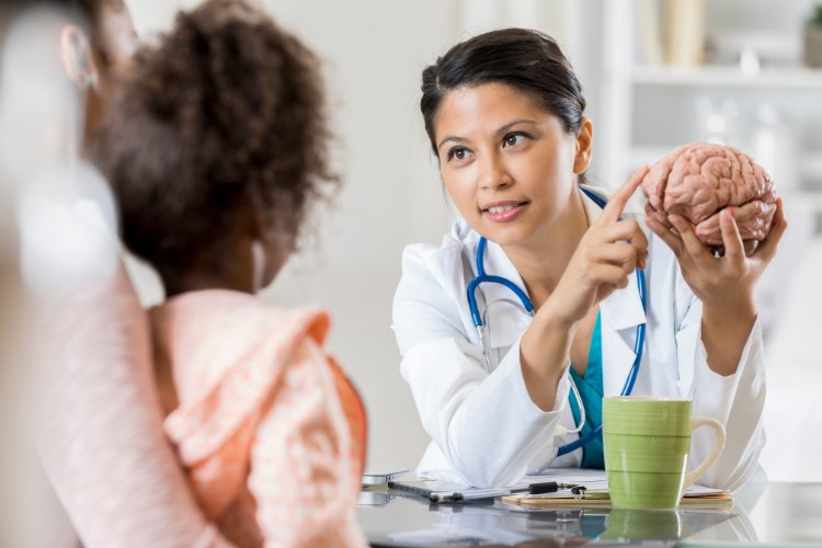 Numero-De-Pessoas-De-Pessoas-Neurodivergentes-No-Brasil-3 Médica de pele morena com imitação de cérebro na mão, mostrando para a mãe e filha