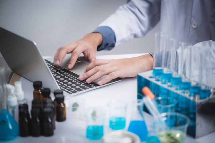 Numero-De-Pessoas-De-Pessoas-Neurodivergentes-No-Brasil-5 Pessoa de pele clara usando jaleco, mexendo no computador, com tubos de ensaio ao lado