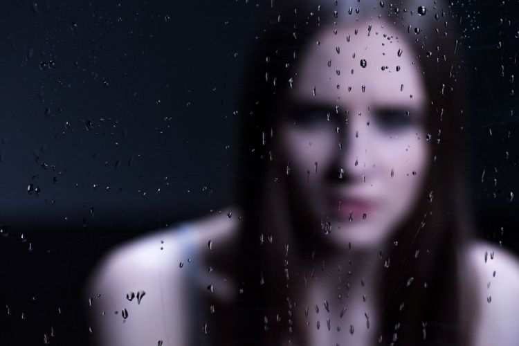 Inseguranca-Ciume-Psicanalise-3 Uma mulher desfocada aparece atrás de um vidro com gotas de chuva, transmitindo melancolia. A imagem simboliza emoções complexas como o ciúme, frequentemente analisadas na psicanálise, que explora inseguranças e sentimentos refletidos em situações de vulnerabilidade emocional