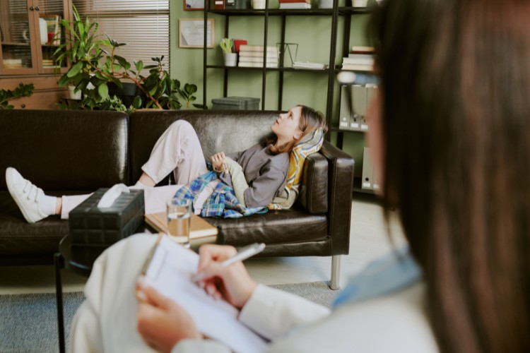 Dinamicas-Familiares-2 Mulher de pele clara na terapia, deitada em sofá preto enquanto outra mulher faz anotações