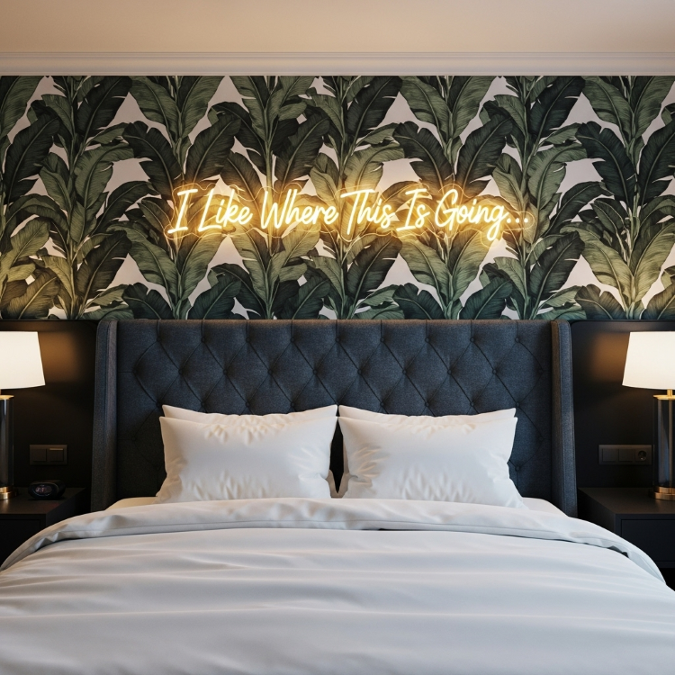 Letreiro-Neon-13 Uma cama com cabeceira estofada azul-marinho. Acima dela, um letreiro de neon dourado com a frase "I Like Where This Is Going.." em uma parede com papel de parede de folhas tropicais verdes e brancas. Duas lâmpadas de mesa estão ao lado da cama.