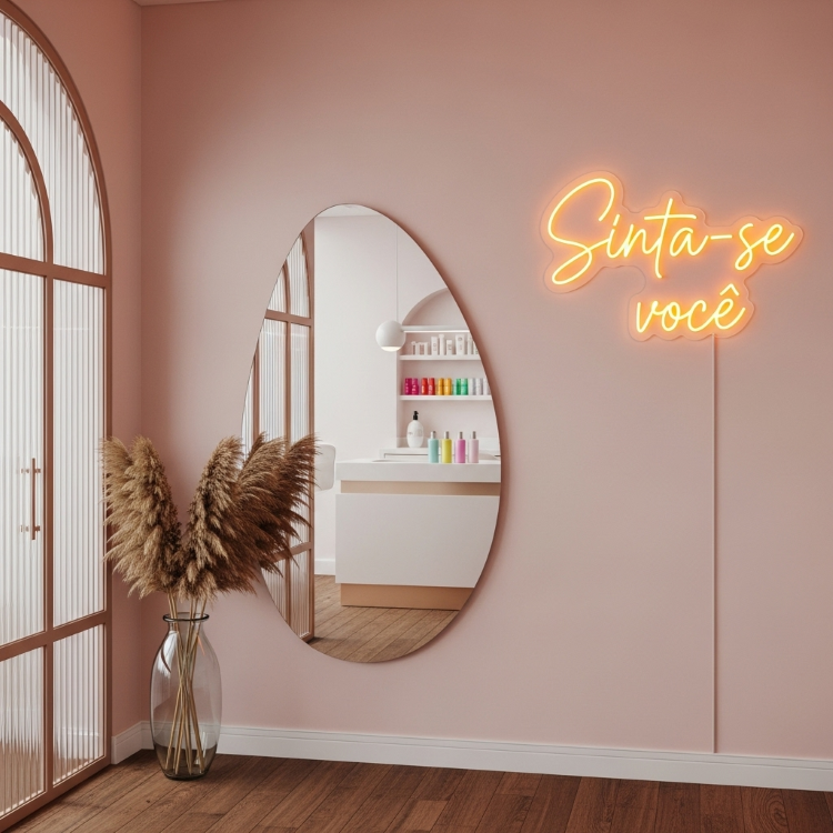 Letreiro-Neon-10 Uma luz de neon laranja com a frase "Sinta-se você" em uma parede rosa. Abaixo, um espelho oval reflete uma bancada branca com potes coloridos e um vaso de vidro com hastes secas de pluma.