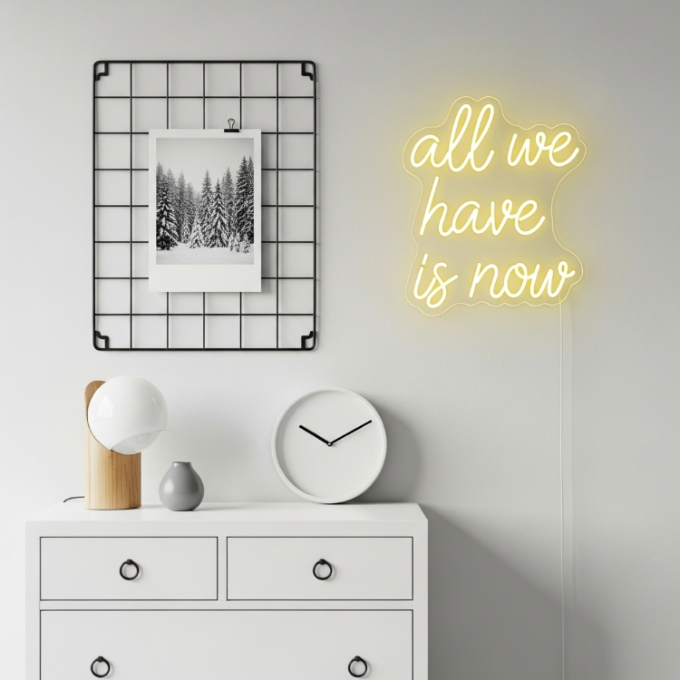 Letreiro-Neon-7 Uma mesa branca com gavetas. Sobre ela, há uma luminária de madeira, um relógio branco, um vaso cinza e, na parede, um neon amarelo diz "all we have is now" ao lado de um quadro de fotos.
