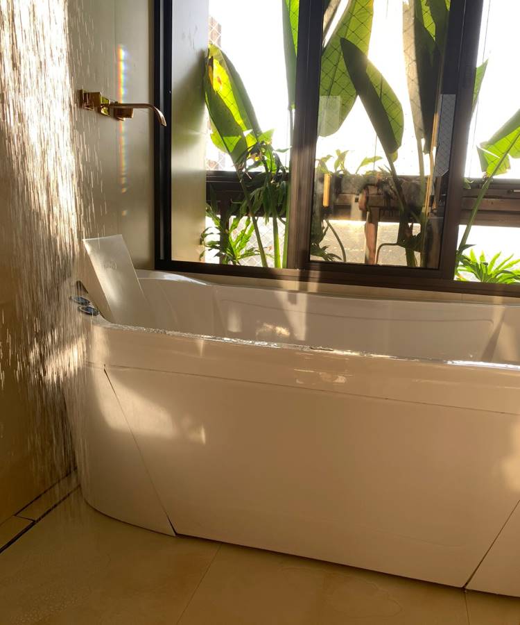 Pretty-Jet-3 Banheira moderna e retangular em um banheiro com grandes janelas, mostrando plantas verdes do lado de fora. A luz do sol da tarde entra pela janela, criando um efeito brilhante na água.