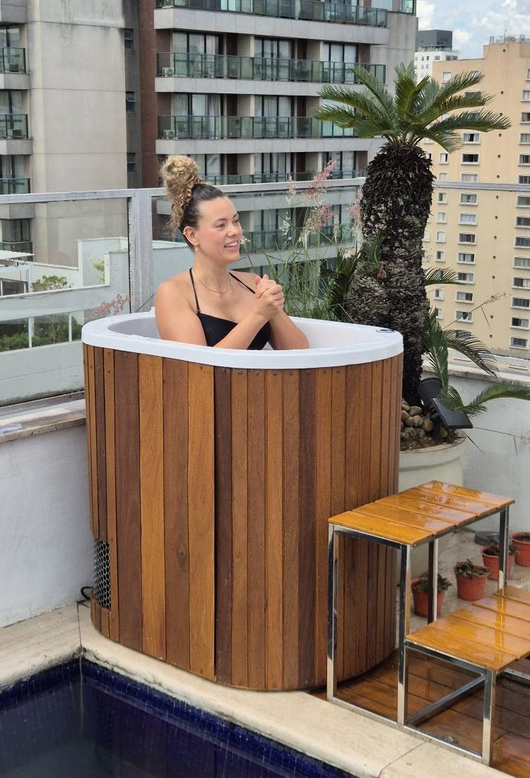 Pretty-Jet-2-Rotated-1 Mulher sorridente relaxando em uma banheira de ofurô de madeira em uma cobertura. A vista ao fundo mostra prédios e uma piscina. Ela está usando um biquíni preto e tem o cabelo preso.
