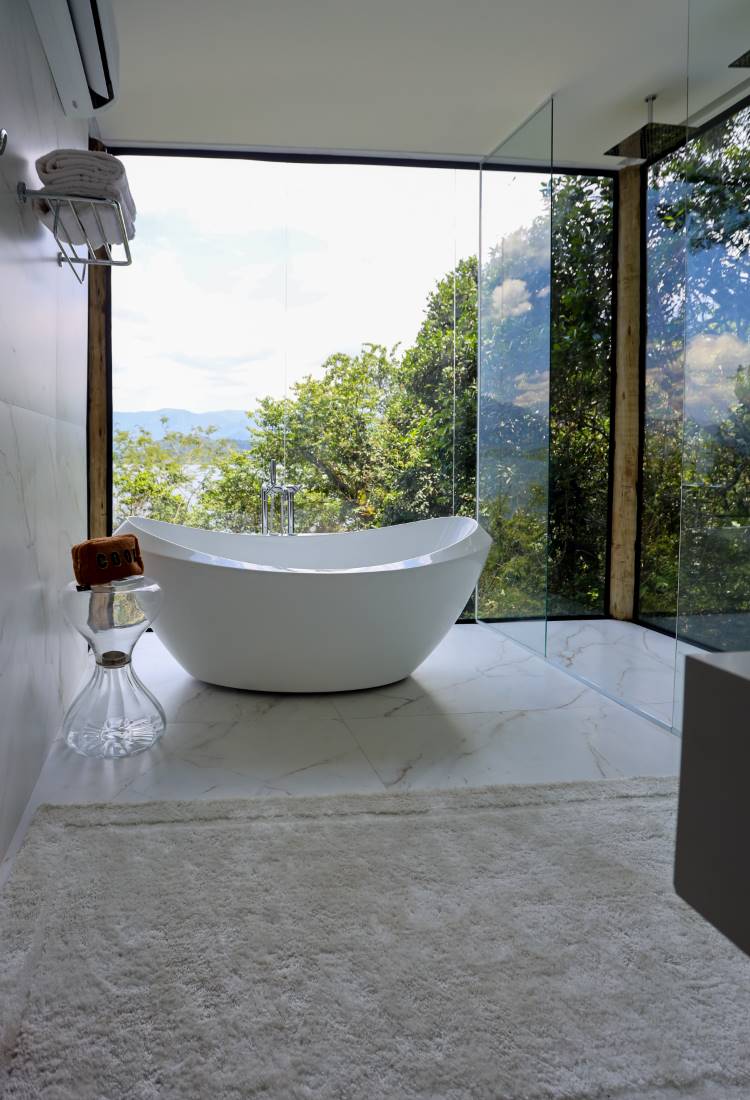 Pretty-Jet-1 Banheira moderna e branca em um banheiro minimalista, com grandes janelas que oferecem uma vista deslumbrante da natureza e do mar. O piso e as paredes são de mármore claro.