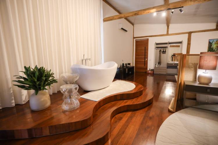 Pretty-Jet-5 Banheira branca moderna em um quarto rústico e aconchegante. A banheira fica em um tablado de madeira curvilíneo, com teto de vigas expostas e uma grande janela com cortina clara.
