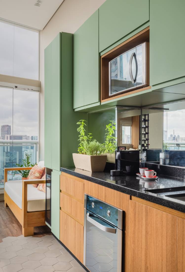 Estudio-77Mc2B2-8 Cozinha moderna com armários superiores em verde-claro e inferiores em madeira. Micro-ondas embutido, bancada preta com planta e máquina de café. Piso de ladrilhos hexagonais brancos na área da cozinha e madeira na varanda. Ao fundo, vista de uma cidade e um sofá branco.