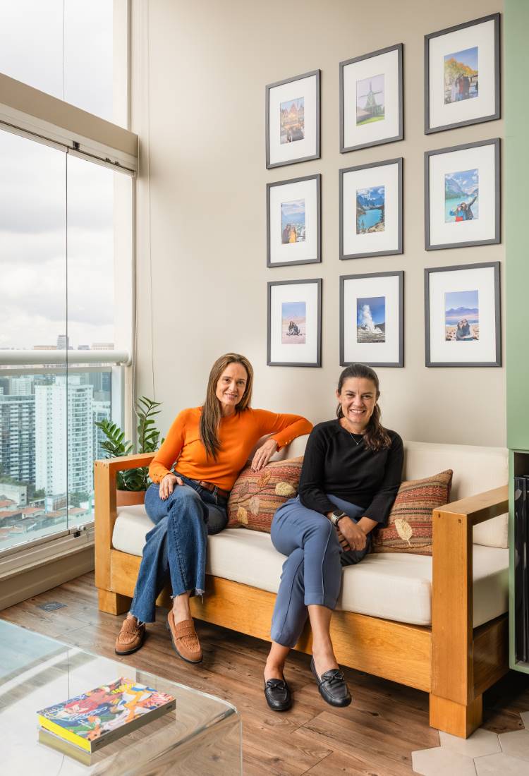 Estudio-77Mc2B2-13 Duas mulheres, sentadas em um sofá de madeira e almofadas estampadas, posam para a foto. A mulher da esquerda veste blusa laranja, a da direita, preta. Atrás delas, uma parede com galeria de nove quadros. Na frente, uma mesa de centro de vidro.