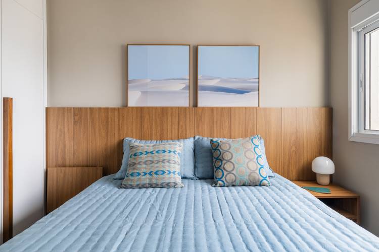 Quarto de casal em um estúdio de 77 m². A imagem mostra uma cama com cabeceira de madeira, roupa de cama azul e duas almofadas estampadas. Na parede, dois quadros com paisagem de dunas. Ao lado, um criado-mudo com abajur branco.