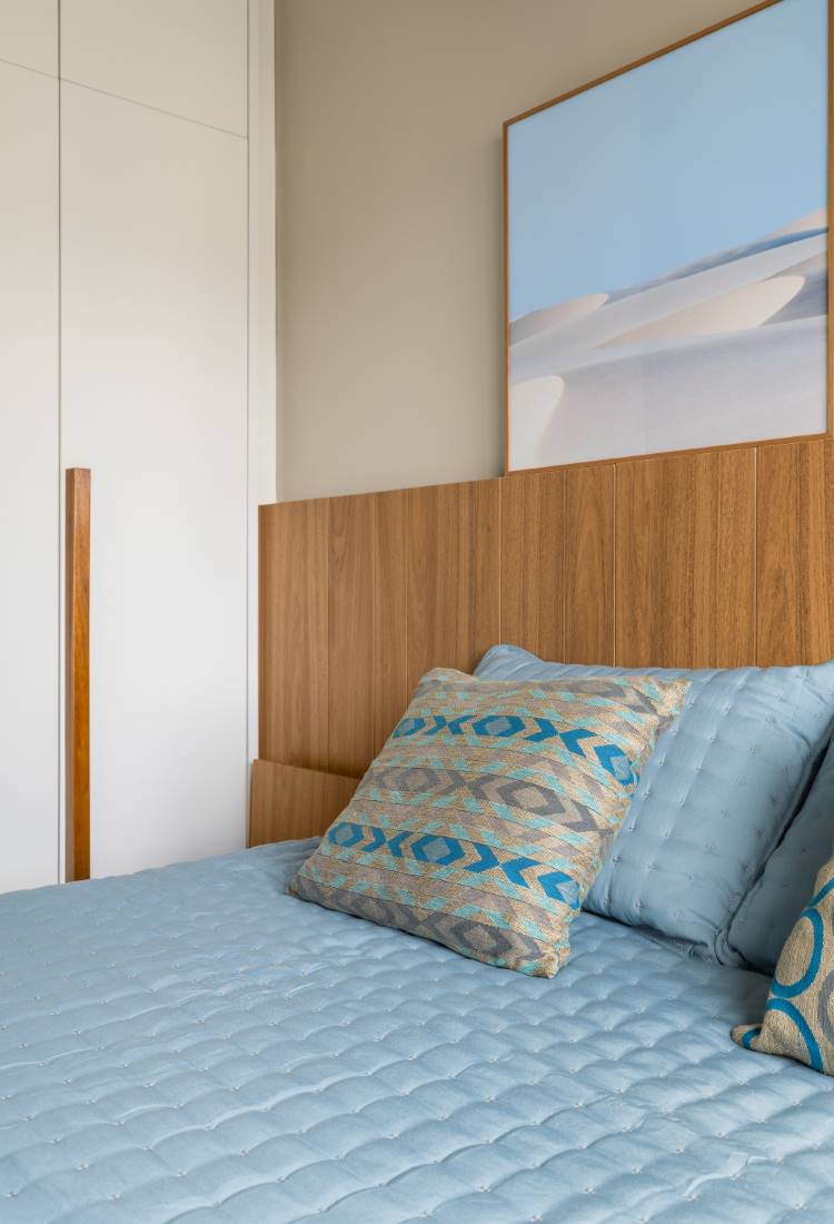 Quarto de um estúdio de 77 m². A imagem mostra uma cama com cabeceira de madeira e roupa de cama azul. No centro, uma almofada decorativa de estampa geométrica. Na parede, um quadro com a paisagem dos lençóis. Ao lado, um guarda-roupa branco.