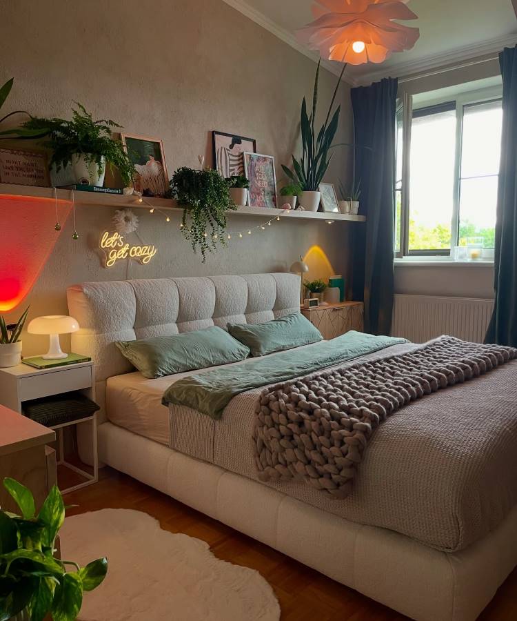 Plantas-Pendentes-13 Uma cama queen size com uma cabeceira estofada e roupa de cama verde e cinza. Prateleiras acima exibem plantas, quadros e luzes de neon. O quarto aconchegante possui iluminação suave e plantas em vasos, criando um ambiente tranquilo.