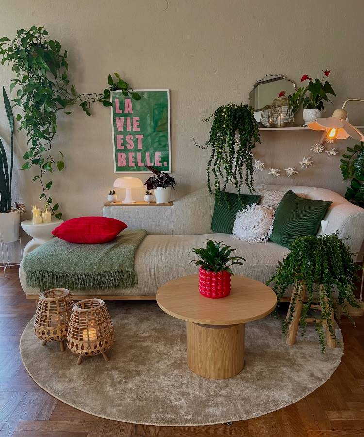 Plantas-Pendentes-11 Uma sala de estar com sofá bege, almofadas verdes e uma manta verde. Plantas em vasos e penduradas decoram o ambiente, com uma mesa de centro redonda de madeira, luminárias de vime e um quadro "La Vie Est Belle" na parede.