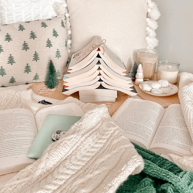 Arvores-De-Natal-Criativas-Livro-1 Livros empilhados em forma de árvore de Natal, com uma fita no topo. Ao redor, há almofadas com estampas de pinheiros, uma vela acesa, copos de bebida, biscoitos em um prato e um suéter tricotado, criando um ambiente aconchegante e festivo.