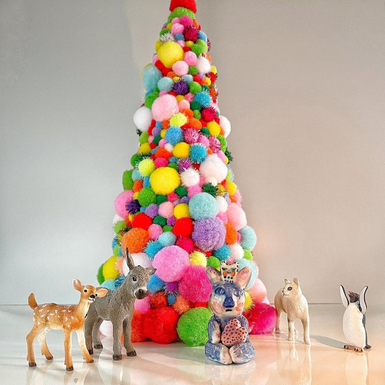 Arvores-De-Natal-Criativas-Pompom-1 Uma árvore de Natal feita de pompons coloridos, cheia de cores vibrantes como rosa, azul, verde e amarelo. Ao redor, cinco pequenas esculturas de animais: dois veados, um coelho, um cavalo e um pinguim, criando uma cena alegre e festiva.