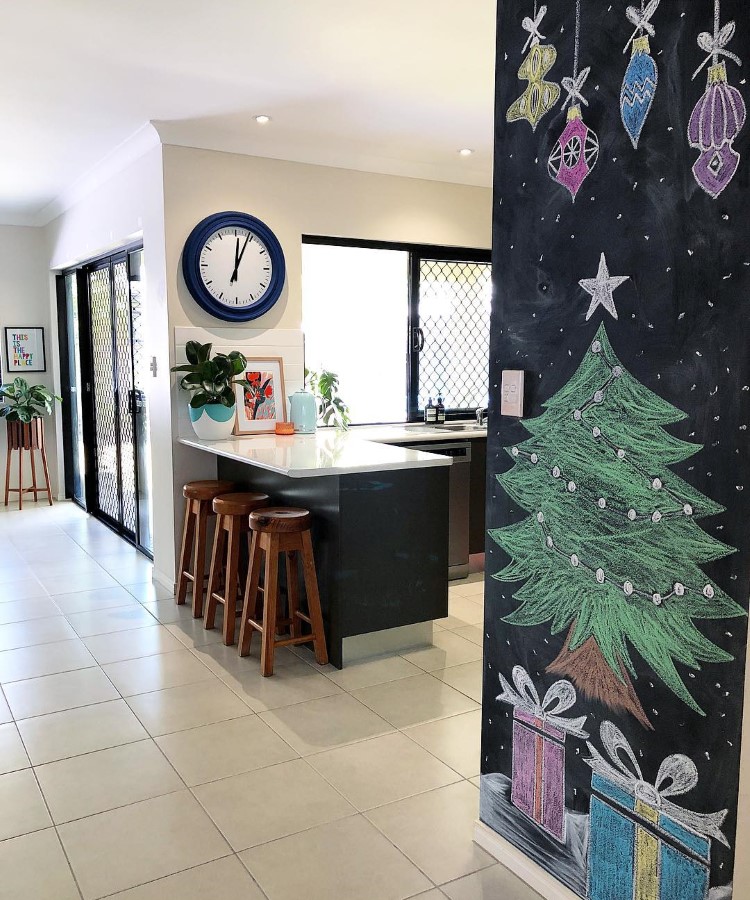 Arvores-De-Natal-Criativas-Quadro-Negro-1 Cozinha moderna com piso claro e balcão preto com banquetas de madeira. Na parede, um relógio azul e quadro. Ao lado, uma lousa com desenho de árvore de Natal e presentes coloridos. Janelas grandes iluminam o ambiente, decorado com plantas.