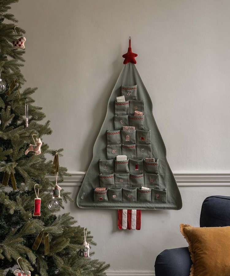 Arvores-De-Natal-Criativas-Tecido-Costura-1 Uma árvore de Natal tradicional está à esquerda, decorada com ornamentos variados. Na parede, um calendário do advento em forma de árvore, com bolsos numerados e uma estrela vermelha no topo. O ambiente é aconchegante, com um sofá azul e almofadas.