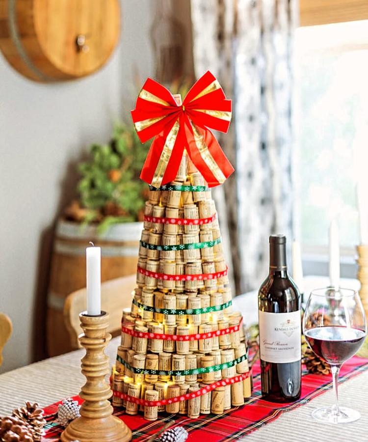 Arvores-De-Natal-Criativas-Rolha-1 Árvore de Natal feita de rolhas de vinho, decorada com fitas vermelhas e verdes, e coroada com um grande laço vermelho. Está sobre uma mesa com toalha xadrez, ao lado de uma garrafa de vinho e uma taça cheia. Ambiente acolhedor e festivo.
