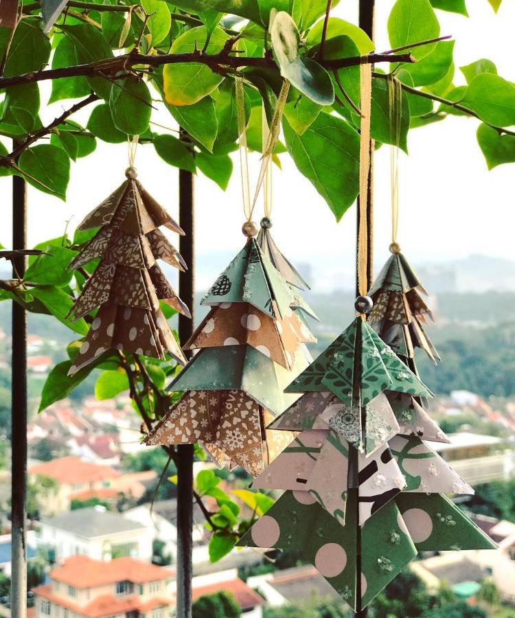 Arvores-De-Natal-Criativas-Origami-1-1 Pequenas árvores de Natal de papel penduradas em ramos de uma planta. Feitas com dobraduras de origami, exibem padrões variados em tons de verde, marrom e bege. Ao fundo, uma vista de uma cidade com telhados vermelhos e vegetação exuberante.