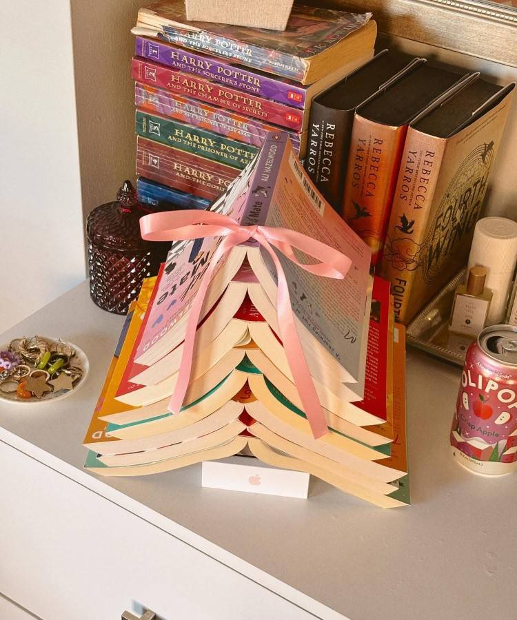 Arvores-De-Natal-Criativas-Livros-1-1 Uma árvore de Natal feita com livros abertos em formato de triângulo, decorada com uma fita rosa no topo. Ao lado, uma pilha de livros "Harry Potter" e outros volumes. Sobre a mesa, itens decorativos e uma lata de bebida.