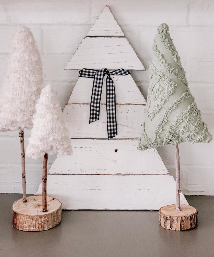 Arvores-De-Natal-Criativas-Pallet-1-1 Três árvores decorativas estão sobre uma superfície. Duas são feitas de tecido, uma branca e outra verde, com texturas diferentes, fixadas em troncos de madeira. Ao fundo, uma árvore de madeira branca com laço xadrez preto e branco, encostada em uma parede clara.