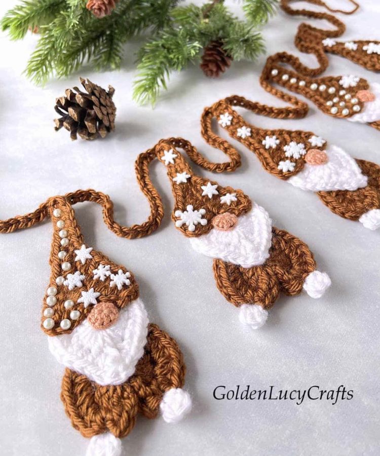 Croche-Natal Adoráveis gnomos de crochê, decorados com pérolas e flocos de neve, estão alinhados sobre uma superfície clara. Ao fundo, um ramo de pinheiro e uma pinha complementam o tema natalino artesanal.