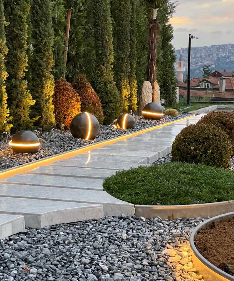 Pedras-Para-Jardim-Ideias-4 Caminho de mármore iluminado por lâmpadas esféricas modernas com detalhes dourados, cercado por brita cinza e jardins com grama e arbustos podados. Árvores altas proporcionam privacidade, enquanto ao fundo se vê um cenário urbano ao entardecer.