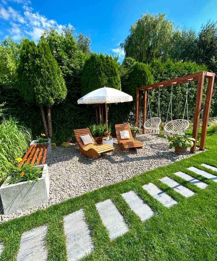 Pedras-Para-Jardim-Ideias-2 Jardim com chão de brita cercado por grama verde. Duas espreguiçadeiras de madeira com almofadas brancas estão próximas a um guarda-sol. Uma estrutura de madeira suporta balanços de macramê. Plantas e árvores criam um ambiente natural e aconchegante.