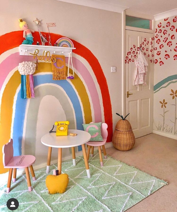Casa-Decorada-Lgbtqia-4 Quarto infantil alegre com parede pintada de arco-íris, mesa branca com cadeiras rosa e verde, telefone retrô amarelo, prateleira decorada com brinquedos, almofadas coloridas, cesto em formato de abacaxi, tapete verde geométrico e detalhes lúdicos.