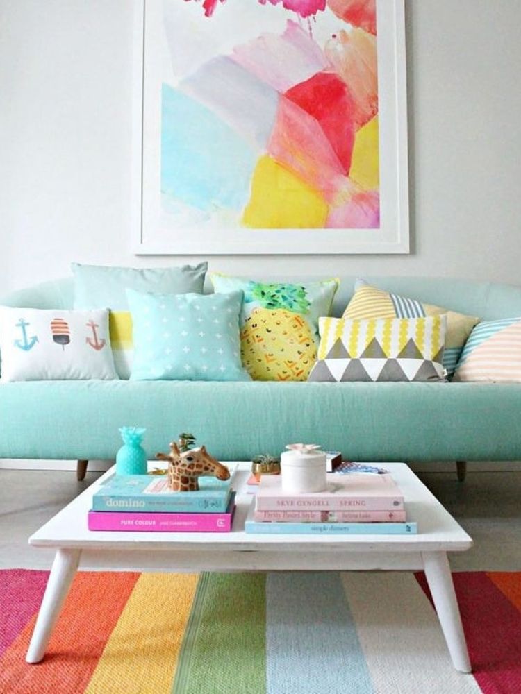 Decoracao-Lgbt-Sala-1 Sala de estar colorida com sofá azul, quadro colorido axima do sofá, almofadas em tons de azul, amarelo e cinza, mesa de centro branca com livros coloridos e tapete nas cores do arco-iris