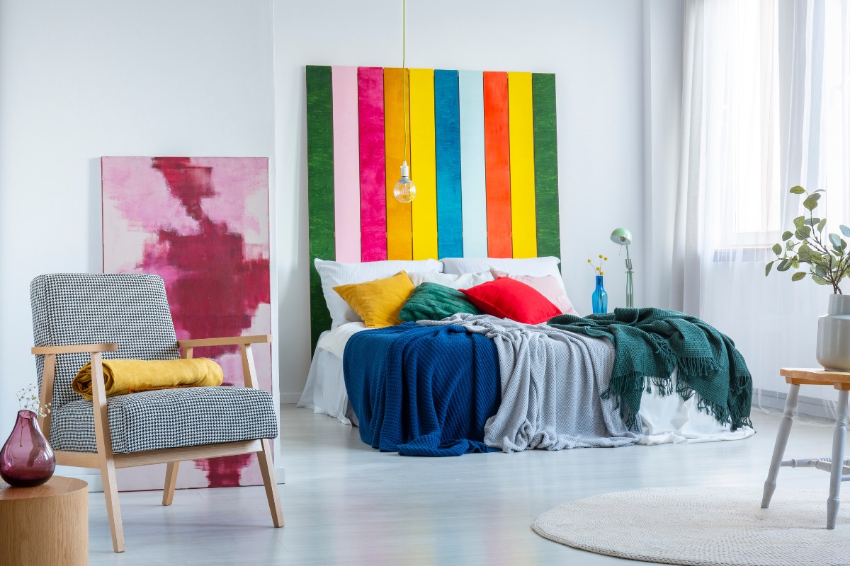 Decoracao-Lgbt-Destacada Foto de sala com quadro nas cores da bandeira LGBT, sofá branco com almofadas coloridas e cobertores, tapete bege, poltrona cinza e amarela e quadro rosa