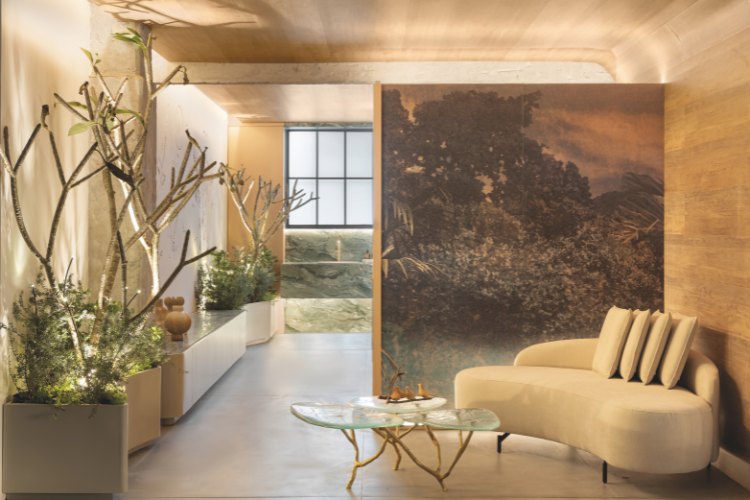 Casacor-Sao-Paulo-2025-14 Hall de banheiro com sofá bege, mesa de centro de vidro, plantas, painel de paisagem e pias ao fundo