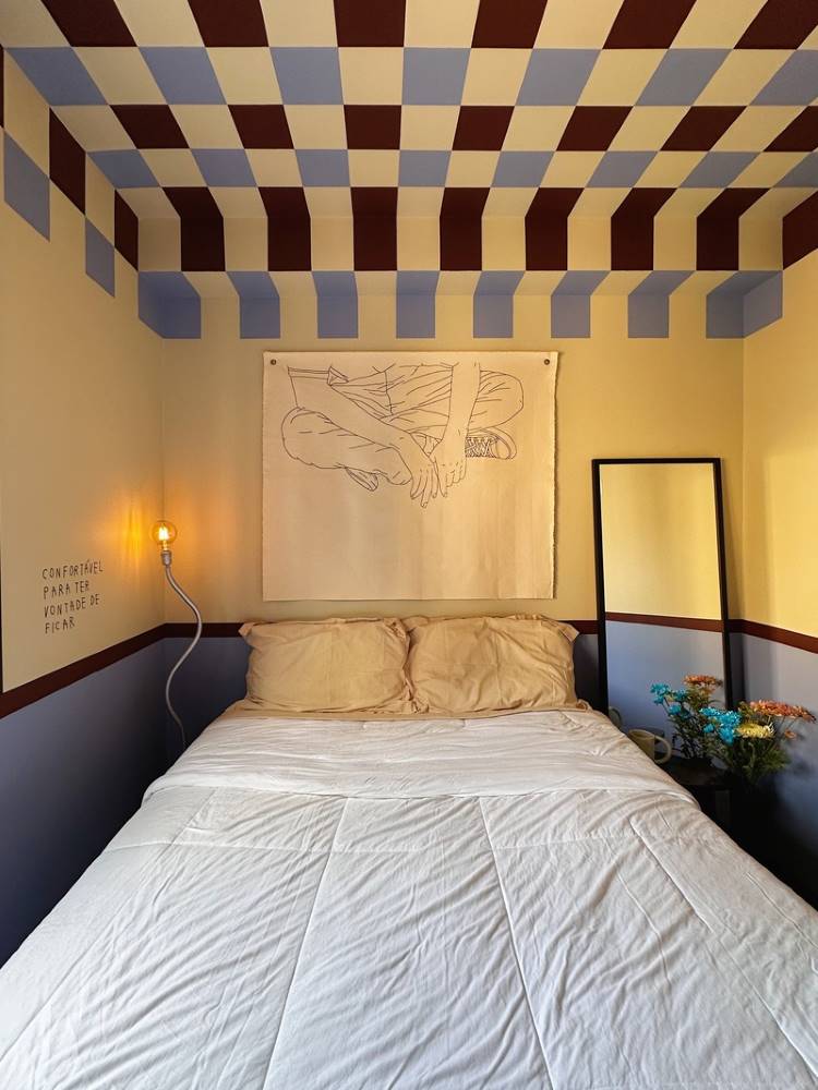 Teto-Colorido-1 Esta é uma imagem de um quarto com cama de casal e roupa de cama clara. O teto e as paredes superiores têm um padrão geométrico em azul claro e bordô. Um espelho alto e uma luminária de piso com uma lâmpada exposta adicionam detalhes ao ambiente.