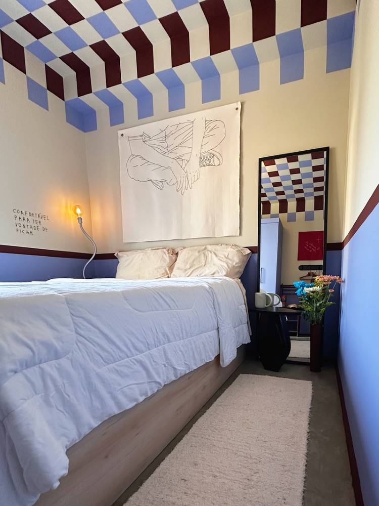 Teto-Colorido-12 Esta imagem mostra um quarto com uma cama de casal e roupa de cama branca. O quarto possui um teto e paredes com um padrão geométrico em tons de azul e bordô. Um espelho alto reflete o padrão e há uma arte abstrata na parede.