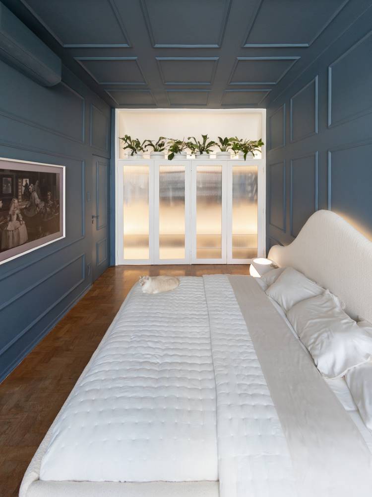 Teto-Colorido-3 Esta é uma imagem de um quarto elegantemente decorado. Uma cama grande com roupa de cama branca domina o centro, com um gato creme deitado nela. As paredes e o teto são de um azul-petróleo profundo com painéis. Ao fundo, portas de vidro fosco com plantas em cima adicionam um toque de natureza.