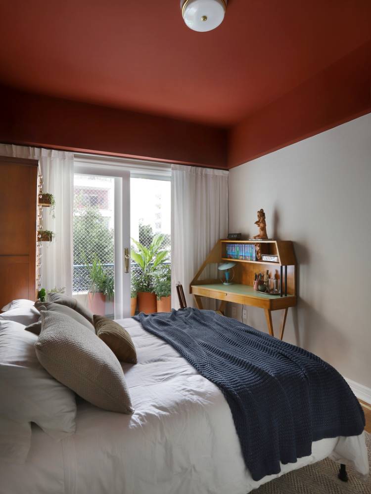 Teto-Colorido-4 Esta imagem mostra um quarto com uma cama de casal e roupa de cama branca, coberta por um cobertor azul-marinho. O teto é pintado de terracota, contrastando com as paredes claras. Uma escrivaninha de madeira está ao lado da cama, e uma porta de vidro se abre para uma varanda com plantas.