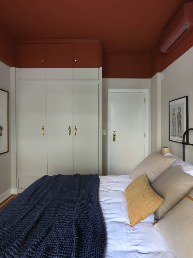 Teto-Colorido-5 Esta imagem mostra um quarto com um teto colorido em tom terracota, que contrasta com as paredes claras. Uma cama grande com roupa de cama branca e um cobertor azul-marinho domina a cena. Armários brancos com puxadores dourados se estendem até o teto, e uma porta branca é visível ao lado.